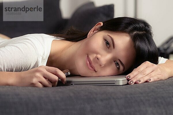 Attraktive junge Frau  die ihren Kopf auf einen Laptop stützt  während sie sich auf dem Sofa ausruht und der Kamera ein charmantes  freundliches Lächeln schenkt