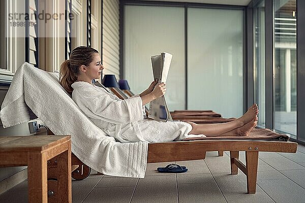 Junge Frau  die sich nach einer Verwöhnbehandlung in einem Spa entspannt  auf einer Liege auf einer Terrasse sitzt und in einem weißen Bademantel eine Zeitung liest