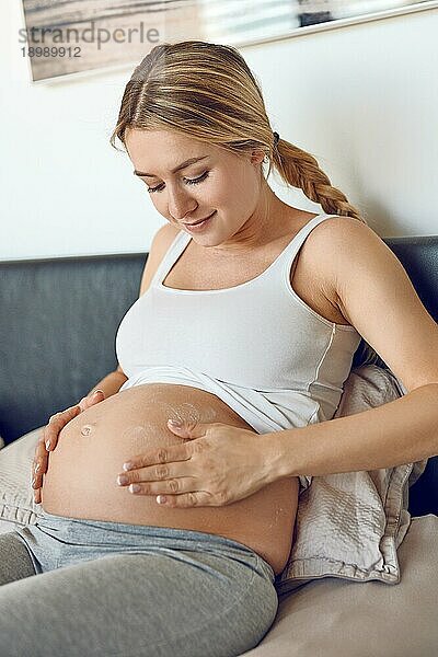 Junge schwangere Frau  die ihren Bauch mit Feuchtigkeitscreme einreibt  um ihre Haut mit Feuchtigkeit zu versorgen und die Gefahr von Schwangerschaftsstreifen nach der Geburt zu verringern