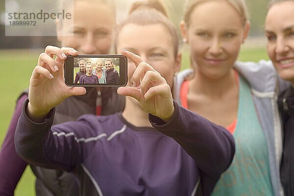 Halbkörperaufnahme von vier glücklichen Frauen  die ein Selfie mit ihrem Smartphone machen  nachdem sie eine körperliche Übung im Freien im Park gemacht haben