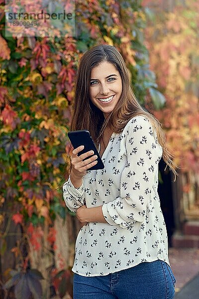 Junge Frau vor buntem Herbstlaub an einer mit Schlingpflanzen bedeckten Mauer  die ihr Handy hält und mit einem warmen  freundlichen Lächeln in die Kamera blickt