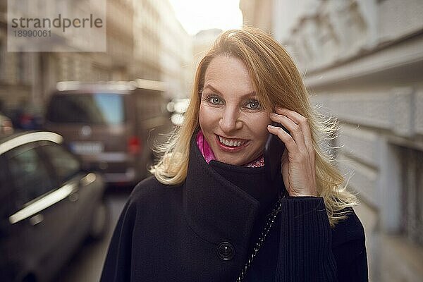Hübsche  lebhafte Frau  die auf einem Mobiltelefon plaudert  während sie eine belebte Straße entlangläuft und fröhlich in die Kamera lächelt