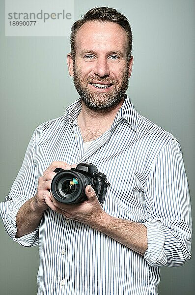 Gutaussehender Fotograf mit einem freundlichen Lächeln  der seine Kamera lachend in die Kamera hält  Oberkörperporträt in entspannter Pose auf grauem Untergrund
