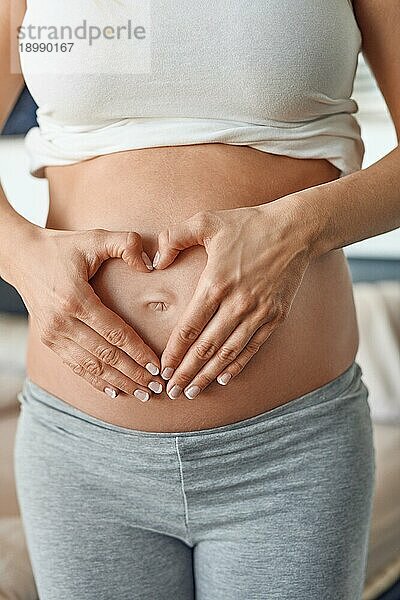 Junge schwangere Frau macht eine Herzgeste mit ihren Händen um ihren nackten Bauchnabel  um ihre Liebe für ihr ungeborenes Kind zu signalisieren