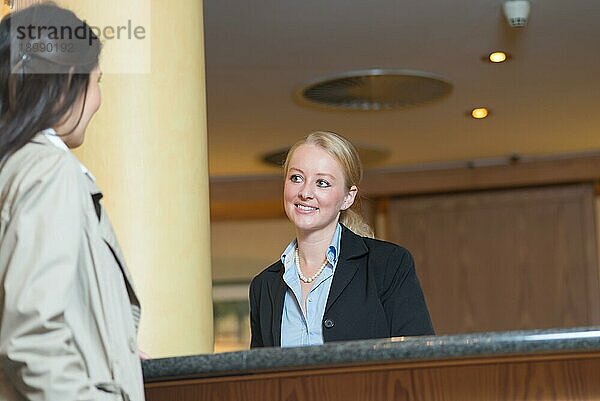 Schöne stilvolle blonde Hotelrezeptionistin steht hinter dem Service Schreibtisch in einer Hotellobby und sieht einen Hotelgast mit einem freundlichen Lächeln an