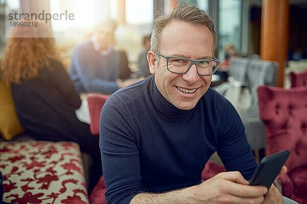 Attraktiver Mann mittleren Alters mit Brille sitzt in einem Restaurant und hält sein Handy in der Hand  während er in die Kamera lächelt