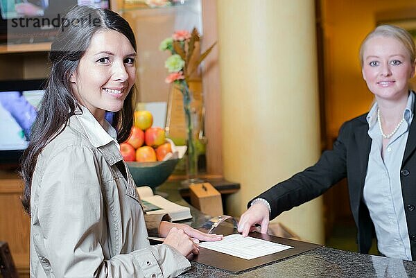 Lächelnder attraktiver junger weiblicher Gast in einer Hotellobby  der sich mit der Rezeptionistin unterhält  als sie bei ihrer Ankunft eincheckt