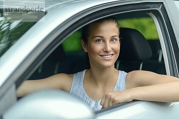 Lächelnde attraktive junge Frau am Steuer eines Autos  die mit einem warmen freundlichen Lächeln aus dem offenen Seitenfenster in die Kamera schaut