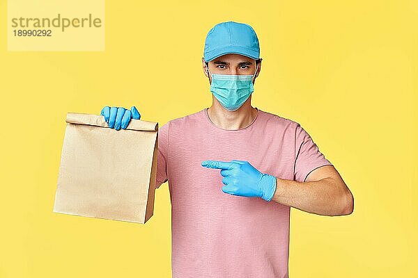 Zusteller  der auf ein Papierpaket mit Lebensmitteln zeigt  trägt Gesichtsmaske und Handschuhe zum Schutz vor dem COVID 19 Coronavirus