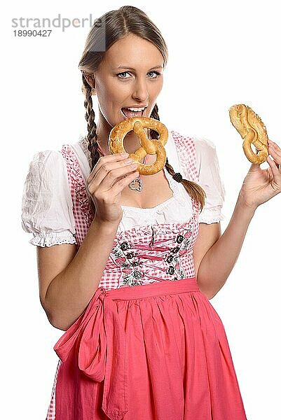 Glückliche attraktive junge deutsche oder bayerische Frau im Dirndl  die zwei Brezeln in der Hand hält und den Mund weit öffnet  um einen Bissen zu nehmen  vor weißem Hintergrund