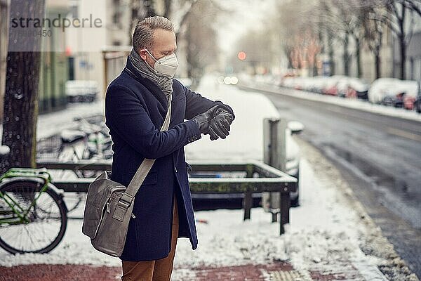 Geschäftsmann mit Umhängetasche und Gesichtsmaske während der Covid 19 Pandemie  der auf einer verschneiten Straße im Winter steht und Zeit auf seiner Armbanduhr überprüft