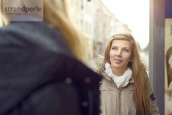 Attraktive junge blonde Frau im Gespräch mit einer Freundin im Freien auf einer belebten städtischen Straße  die lächelt  während sie ihrem Gespräch zuhört