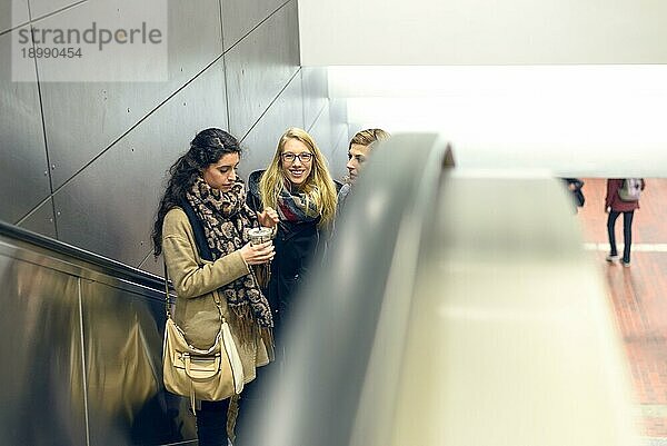 Drei junge erwachsene Frauen in Mantel und Schals fahren auf einer Rolltreppe in einem Gebäude mit seitlichem Kopierraum nach oben