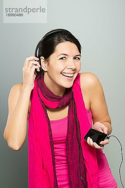 Schöne  lebhafte Frau  die über Kopfhörer  die an ein tragbares elektronisches Speichergerät angeschlossen sind  Musik hört  während sie lachend in die Kamera blickt