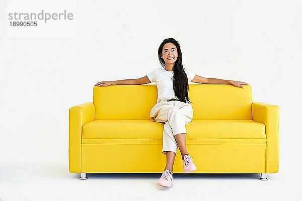 Hübsche lächelnde asiatische Frau entspannen auf gelber Couch auf weißem Hintergrund. Ruhe  Komfort Konzept