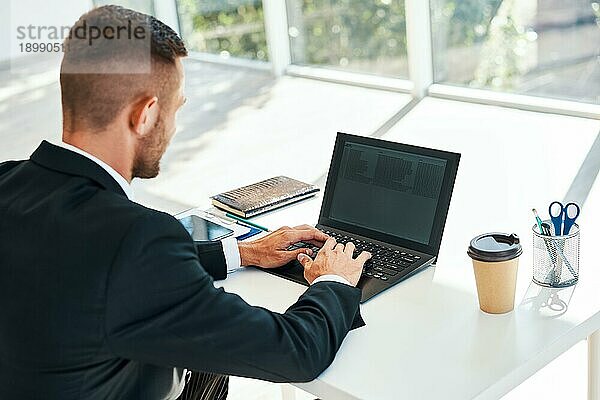 Rückansicht eines Geschäftsmannes  der mit einem Laptop in einem modernen Bürointerieur auf seinem Schreibtisch sitzt. Menschen im Geschäft