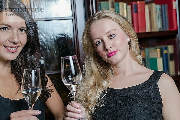 Zwei elegante  kultivierte junge Frauen in stilvoller schwarzer Abendgarderobe feiern zusammen und trinken Champagner in einer Hotelbibliothek oder einem luxuriösen Hausinterieur