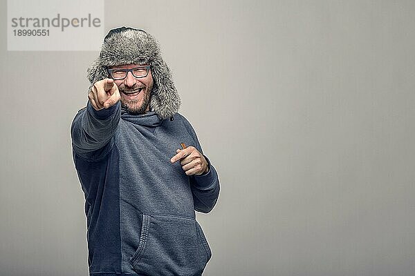 Lachender  jovialer Mann mit Brille und pelziger Wintermütze  der mit spielerischem Ausdruck auf die Kamera zeigt  über grauem Hintergrund mit Kopiervorlage