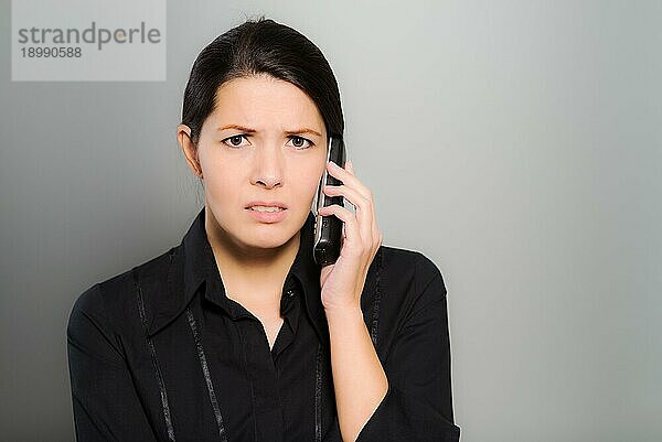 Verwirrte  schöne junge Frau  die mit ihrem Handy telefoniert und mit verwirrtem Blick und Stirnrunzeln in die Kamera schaut  vor einem grauen Studiohintergrund