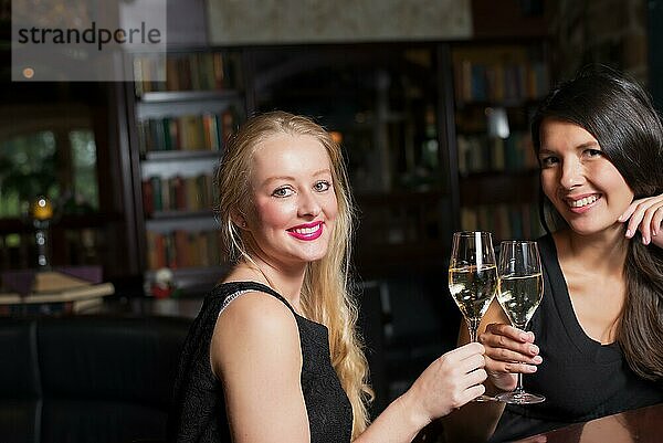 Zwei schöne elegante Frauen Freunde in stilvollen einfachen schwarzen Cocktailkleider Toast einander mit Gläsern von gekühlten Champagner  wie sie auf eine Nacht zusammen feiern