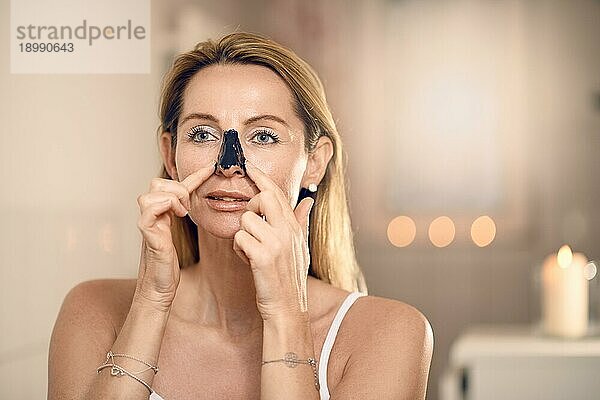 Attraktive blonde Frau mittleren Alters  die eine Anti Aging Gesichtsmaske auf ihre Nase aufträgt  in einem Badezimmer mit brennenden Kerzen in einem Konzept für Schönheit  Hautpflege und den Alterungsprozess mit Kopierraum