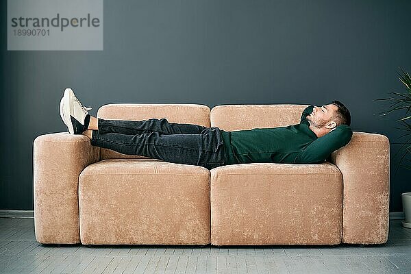 Junger trendiger Mann  der sich allein auf einem Textilsofa zu Hause entspannt. Ruhe Konzept