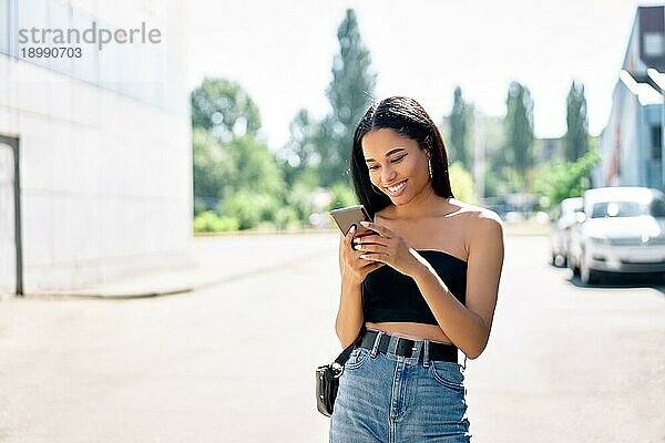 Glückliche lächelnde schwarze Frau  die auf ihr Smartphone schaut  wenn sie in der Stadt spazieren geht. Menschen  Kommunikation Konzept