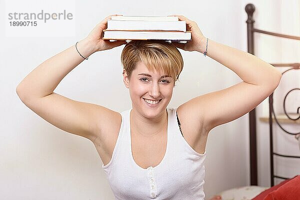 Glückliche  hübsche junge Frau  die eine Pause von ihrem Studium macht und Bücher auf ihrem Kopf balanciert  während sie mit einem warmen  freundlichen Lächeln in die Kamera schaut