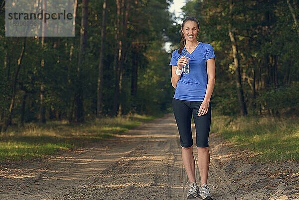 Fitte schlanke junge Frau in Sportkleidung trinkt Wasser in Flaschen  während sie auf einem Waldweg eine Pause einlegt  um während eines Trainingslaufs auf dem Lande neue Flüssigkeit zu sich zu nehmen  Gesundheits und Fitnesskonzept