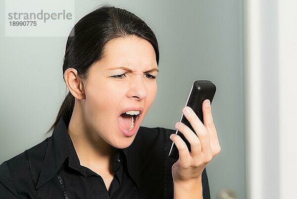 Porträt einer brünetten kaukasischen jungen Frau  die schreit  während sie auf ihr Handy schaut  Symbol für Stress und negative Gefühle  auf grauem Hintergrund