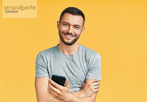 gutaussehender  lächelnder Mann  der ein Mobiltelefon hält und in die Kamera schaut  in einem gelben Studiohintergrund. Kommunikation Konzept