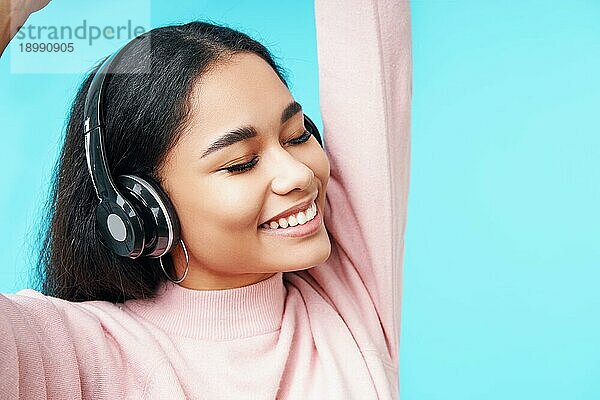 Close up Porträt der hübschen jungen Frau hören Musik mit ihren Kopfhörern über blaün Hintergrund