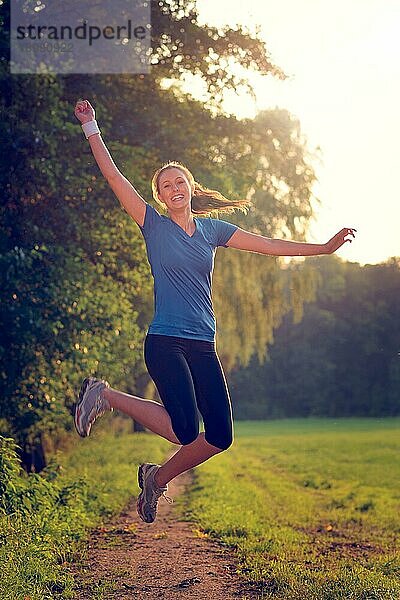 Energetische Frau  die mit einem glücklichen Lächeln voller Vitalität in die Luft springt  während sie während eines körperlichen Trainings auf einem Feldweg läuft