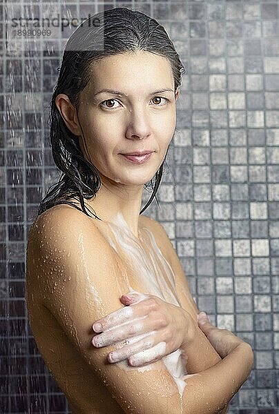 Attraktive Frau  die sich in der Dusche einseift  während sie unter dem Wasserstrahl steht und ihre Brüste mit den Armen bedeckt und in die Kamera schaut  in einem Konzept der Körperpflege