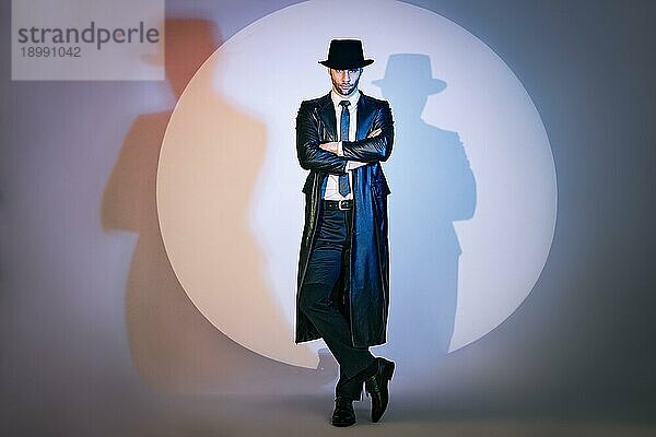 Ganzkörperporträt eines selbstbewussten  gut aussehenden Mannes in schwarzem Mantel und Hut mit verschränkten Armen  der im Studio im Scheinwerferlicht posiert. Noir Film Stil. Privatdetektiv  Spion  Ermittlungskonzept