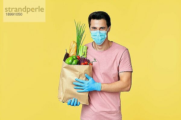 Mann hält eine Papiertüte mit frischem Gemüse in der Hand und trägt Gesichtsmaske und Handschuhe  um sich vor dem Coronavirus Covid19 zu schützen