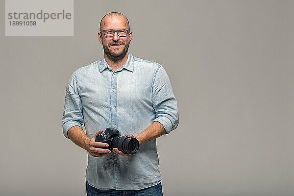 Gutaussehender Fotograf mit einem freundlichen Lächeln  der seine Kamera lachend in die Kamera hält  Oberkörperporträt in entspannter Pose auf grauem Untergrund