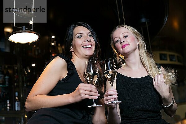 Zwei glückliche Freundinnen  eine Blondine und eine Brünette  stoßen mit Champagner oder Weißwein in einem angesagten Nachtclub an