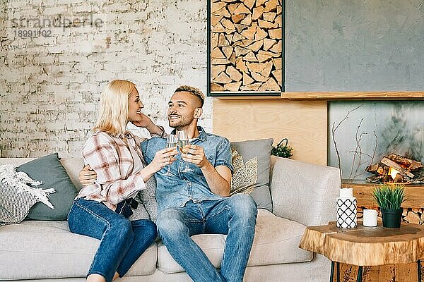 Junges glückliches Paar trinkt Champagner und lächelnd sitzt auf dem Sofa in ihrem modernen Haus. Beziehung  Feier  Dating Konzept
