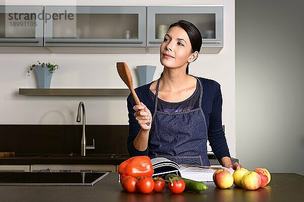 Glückliche hübsche Frau mit Schürze  die eine hölzerne Schöpfkelle hält  während sie ein Rezeptbuch am Tisch mit Obst und Gemüse in der Küche liest