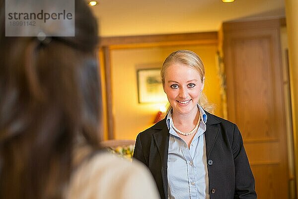 Schöne stilvolle blonde Hotelrezeptionistin steht hinter dem Serviceschalter in einer Hotellobby und schaut mit einem freundlichen Lächeln in die Kamera