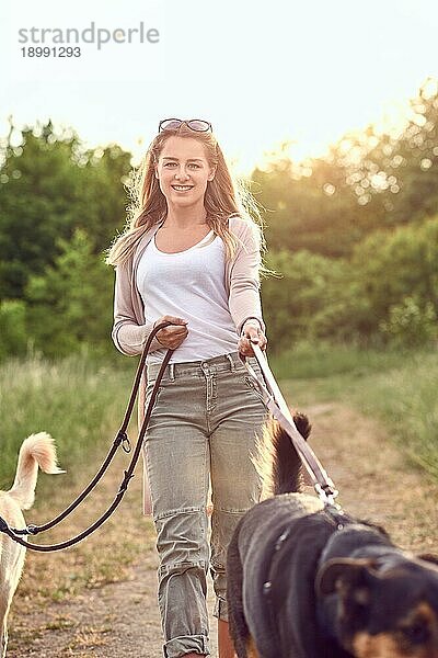 Lächelnde hübsche blonde Frau  die mit ihren beiden Hunden auf einem ländlichen Feldweg spazieren geht  im Gegenlicht der tief über den Baumwipfeln stehenden Sonne  mit Fokus auf die Frau