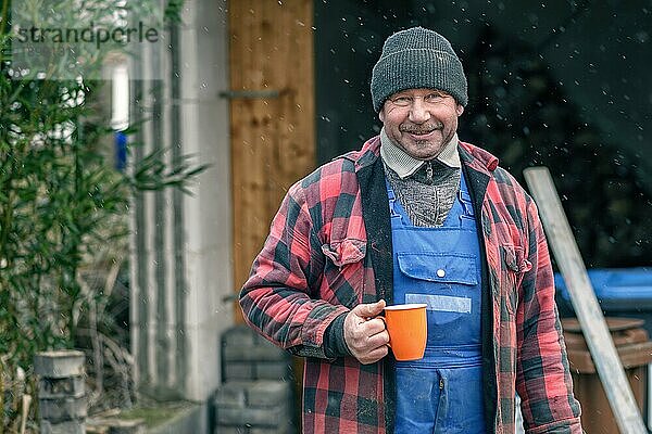 Glücklicher Mann mit gestrickter Wollmütze  Trikot und Jacke  der bei verschneitem Winterwetter im Freien steht und heißen Kaffee aus einem leuchtend orangefarbenen Becher trinkt