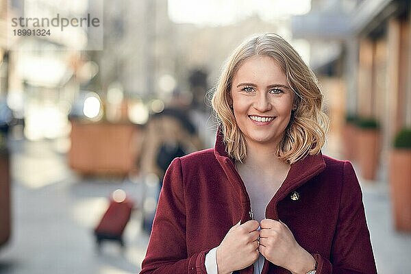 Attraktive junge blonde Frau mit einem freundlichen Lächeln  die einen kastanienbraunen Mantel trägt und durch eine Straße mit Sonnenlicht läuft