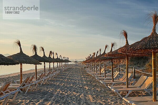 Sonnenaufgang an einem menschenleeren tropischen Resortstrand mit strohgedeckten Sonnenschirmen und Liegestühlen mit Blick auf das ruhige Meer für einen idyllischen Sommerurlaub