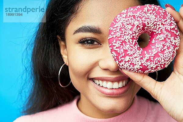 Nahaufnahme Porträt der schönen schwarzen Frau und Donut. Mädchen zeigt Donut vor ihren Augen auf blauem Hintergrund. Leckeres Essen  Diätkonzept