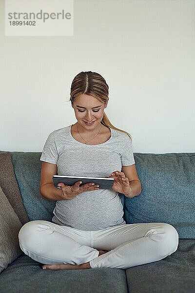 Glückliche schwangere Frau  die sich auf einem Sofa im Schneidersitz entspannt und ein eBook auf einem Tablet liest  mit einem warmen  freundlichen Lächeln