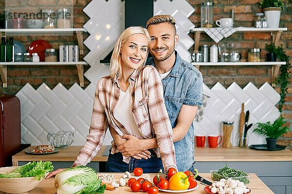 Lächelndes junges Paar kocht zusammen vegetarische Mahlzeit in der Küche zu Hause. Mann umarmt Frau. Gesunder Lebensstil Konzept