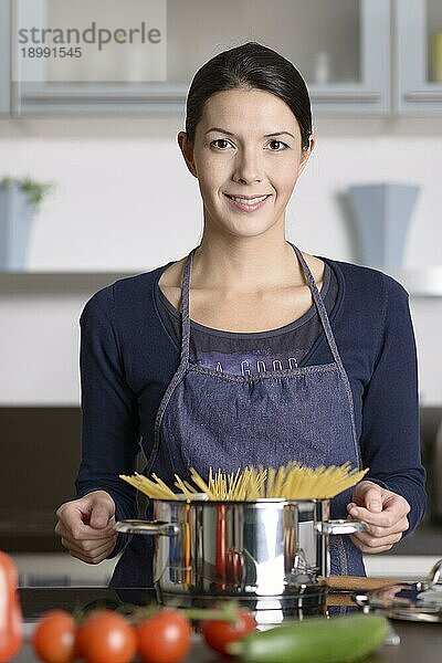 Junge Hausfrau  die eine gesunde italienische Pasta zubereitet  steht am Herd mit einem Topf voller Spaghetti mit frischem Gemüse im Vordergrund und lächelt fröhlich in die Kamera