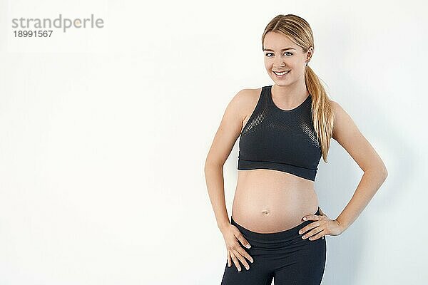 Glückliche  gesunde  junge  schwangere Frau in schwarzer Sportkleidung  die mit den Händen auf den Hüften steht und mit einem strahlenden Lächeln in die Kamera schaut